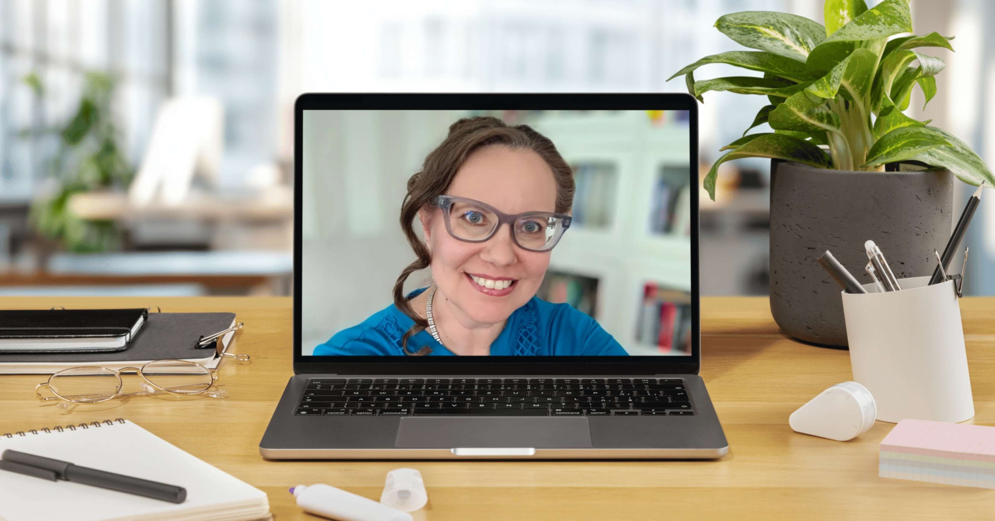 Tietokoneen näytöllä hymyilevä Musiikkikadun Mari etätapaamisessa. Työpöydällä muistivihko, kynä ja kasvi, taustalla vaalea ja valoisa työtila.